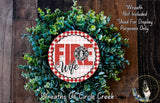 a wreath made of green leaves and a red and white sign that says "FIRE WIFE". The wreath is displayed on a wooden surface, and there is a small dog in the bottom right corner of the image.