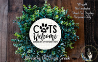 a wreath made of green leaves and a white circular sign with the text "CATS WELCOME HUMANS BY APPOINTMENT ONLY" in the center. The sign is surrounded by a lush wreath, and there is a small dog visible in the bottom right corner of the image.