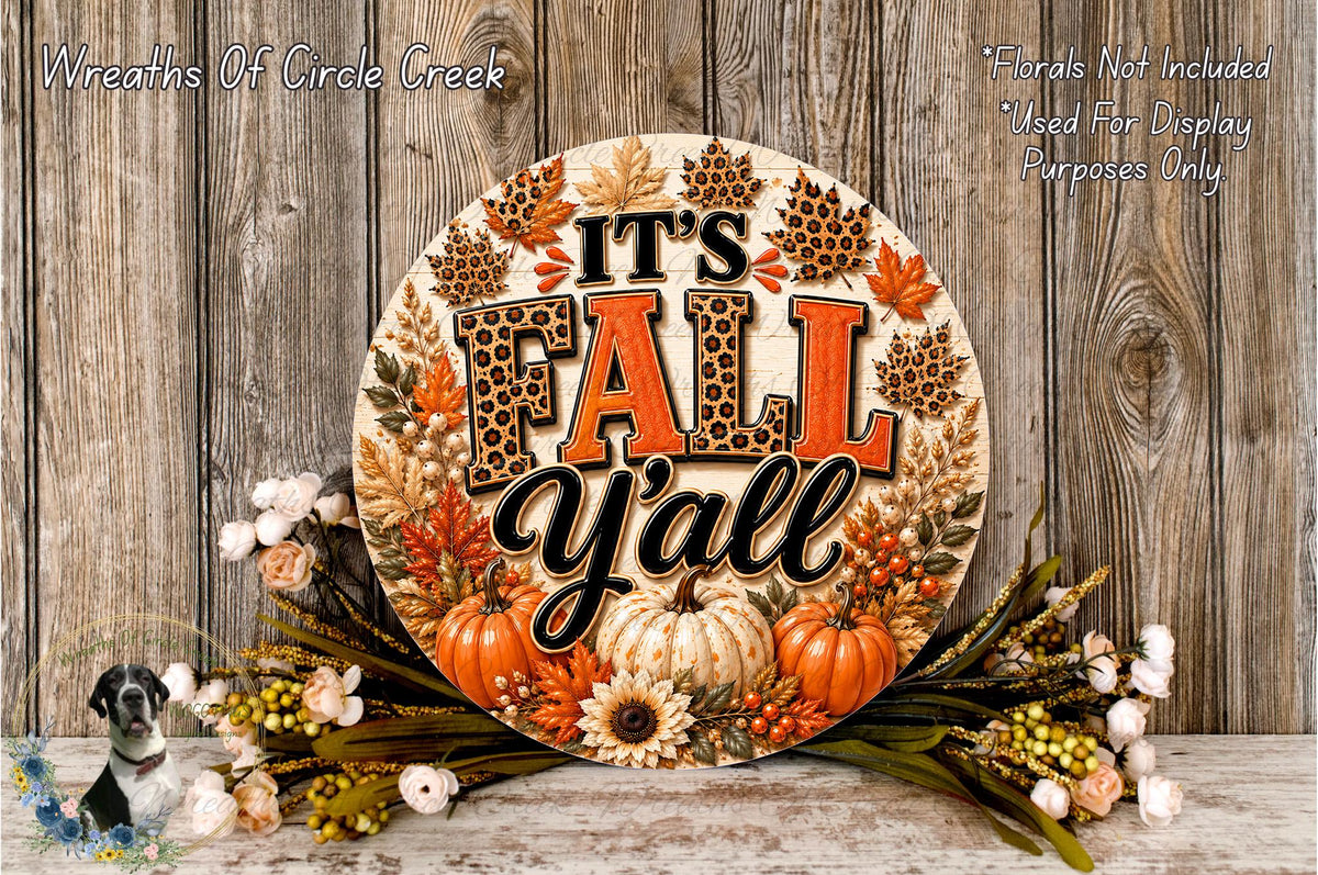 A decorative sign with the text "It's Fall Y'all" surrounded by autumn-themed elements such as leaves, pumpkins, and flowers, placed on a wooden surface.