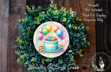 A wreath made of green leaves and colorful balloons, with a birthday cake and presents on top, is displayed on a wooden surface.