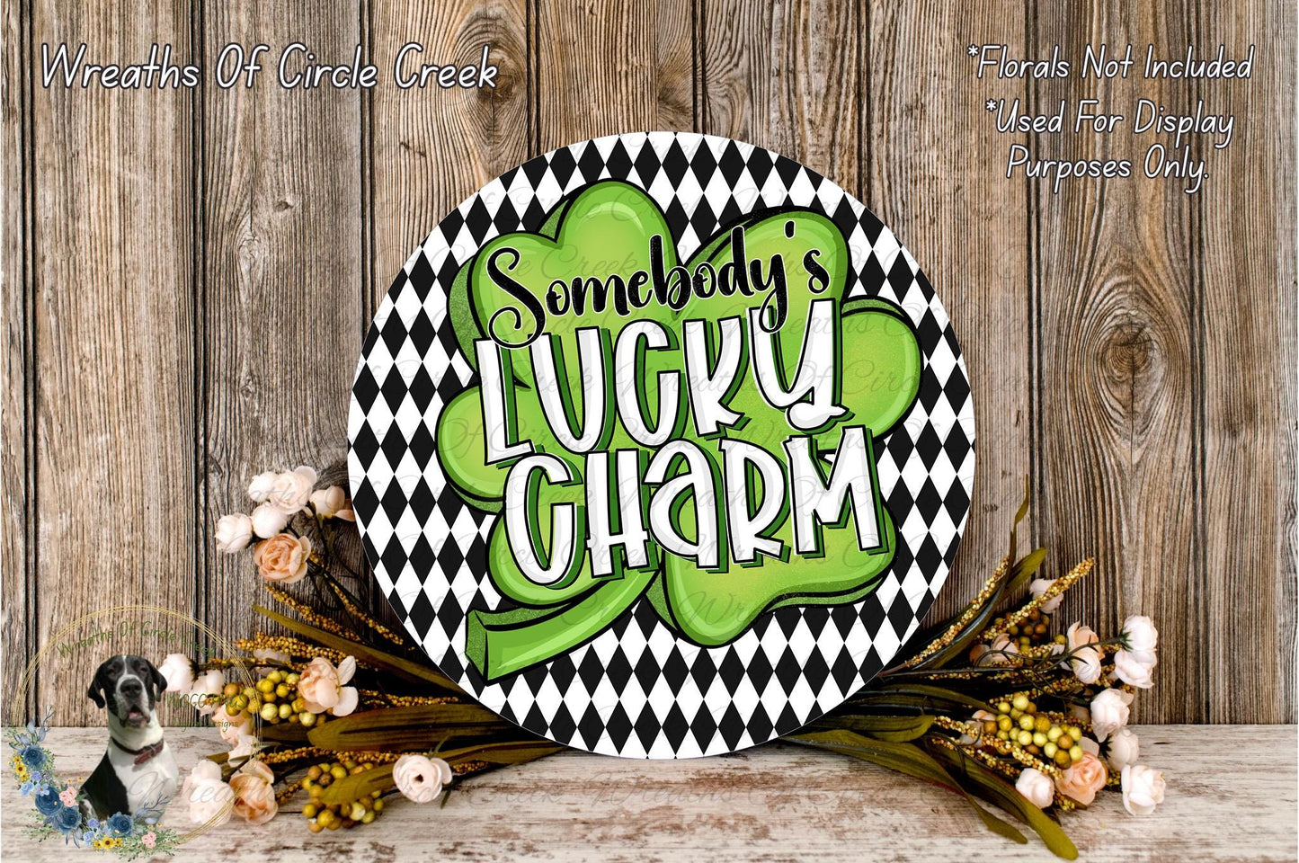 a circular sign with the text "SOMEBODY'S LUCKY SHARM" written on it, surrounded by a black and white checkered pattern and a green shamrock. The sign is placed on a wooden surface, with a wreath of flowers and greenery to the side.