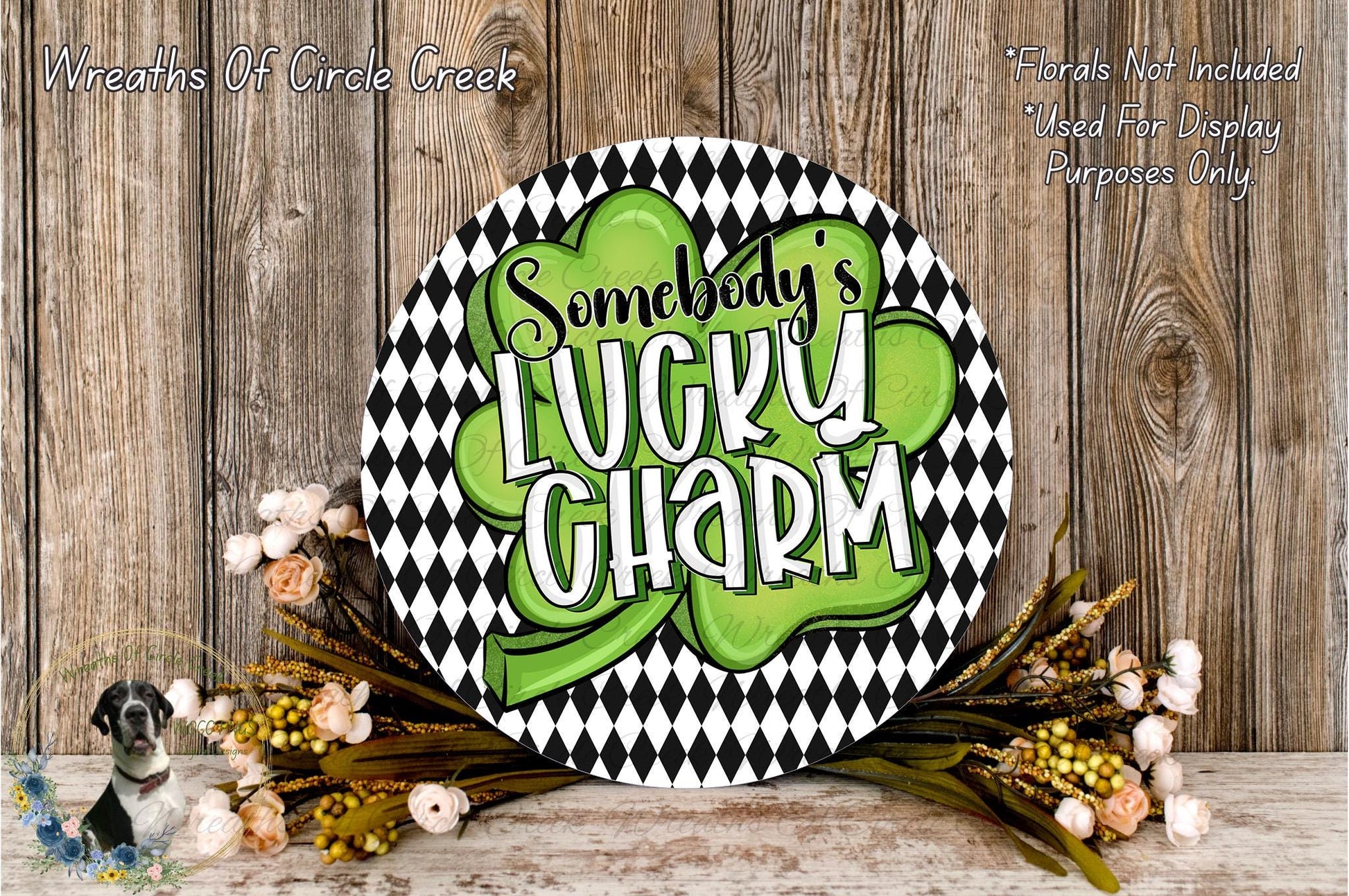 a circular sign with the text "SOMEBODY'S LUCKY SHARM" written on it, surrounded by a black and white checkered pattern and a green shamrock. The sign is placed on a wooden surface, with a wreath of flowers and greenery to the side.