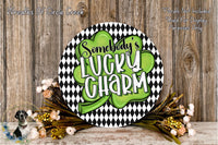 a circular sign with the text "SOMEBODY'S LUCKY SHARM" written on it, surrounded by a black and white checkered pattern and a green shamrock. The sign is placed on a wooden surface, with a wreath of flowers and greenery to the side.