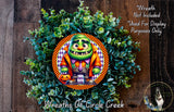 A wreath featuring a cartoon character with a green body, orange and purple accents, and a pumpkin, surrounded by a lush green plant.
