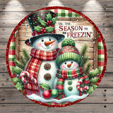 A festive winter scene featuring two snowmen, one larger and one smaller, wearing colorful scarves and hats, surrounded by snow, holly, and other holiday decorations. The text "Tis the SEASON to be FREEZIN'" is displayed above the snowmen, suggesting a seasonal theme.