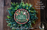A Christmas wreath made of evergreen branches and a red and white checkered border, with a snowman and gingerbread man decoration. The wreath is surrounded by greenery and has a "Merry Christmas" sign in the center.