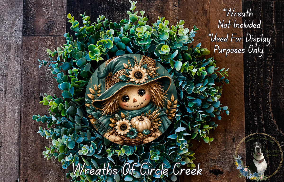 a wreath with a scarecrow design, surrounded by various green foliage and flowers. The wreath is displayed on a wooden surface, and there is a small dog in the bottom right corner of the image.