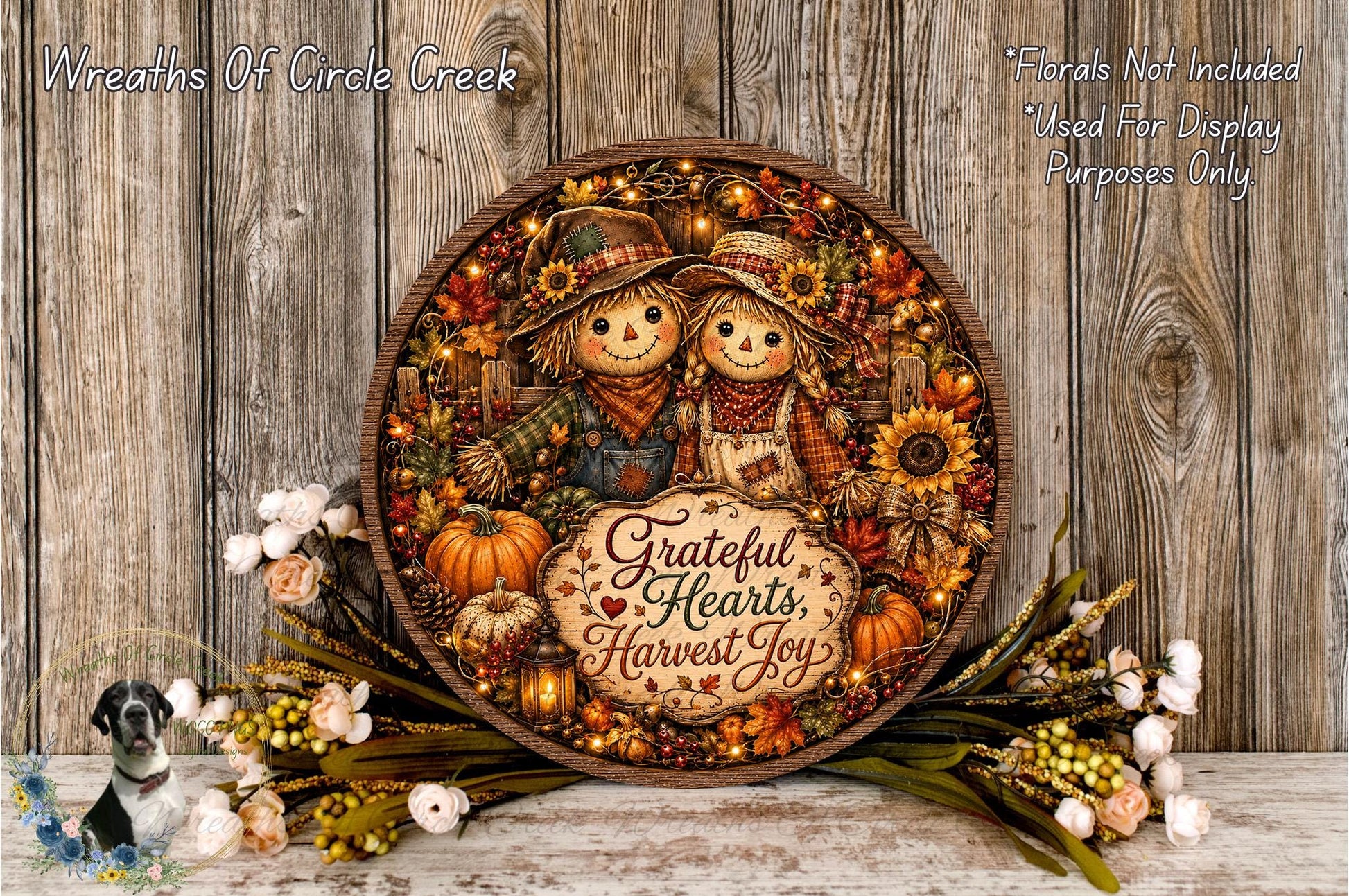 A decorative wreath with a couple of scarecrow figures, surrounded by autumn-themed elements such as pumpkins, sunflowers, and pine cones. The wreath is placed on a wooden surface, and there are flowers and greenery surrounding it.