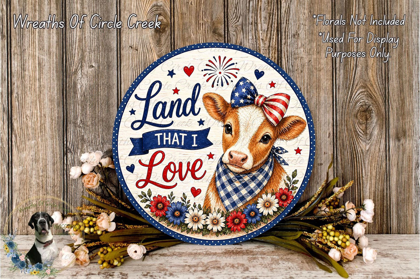 a decorative sign with a cow wearing a patriotic bow, surrounded by flowers and placed on a wooden surface.