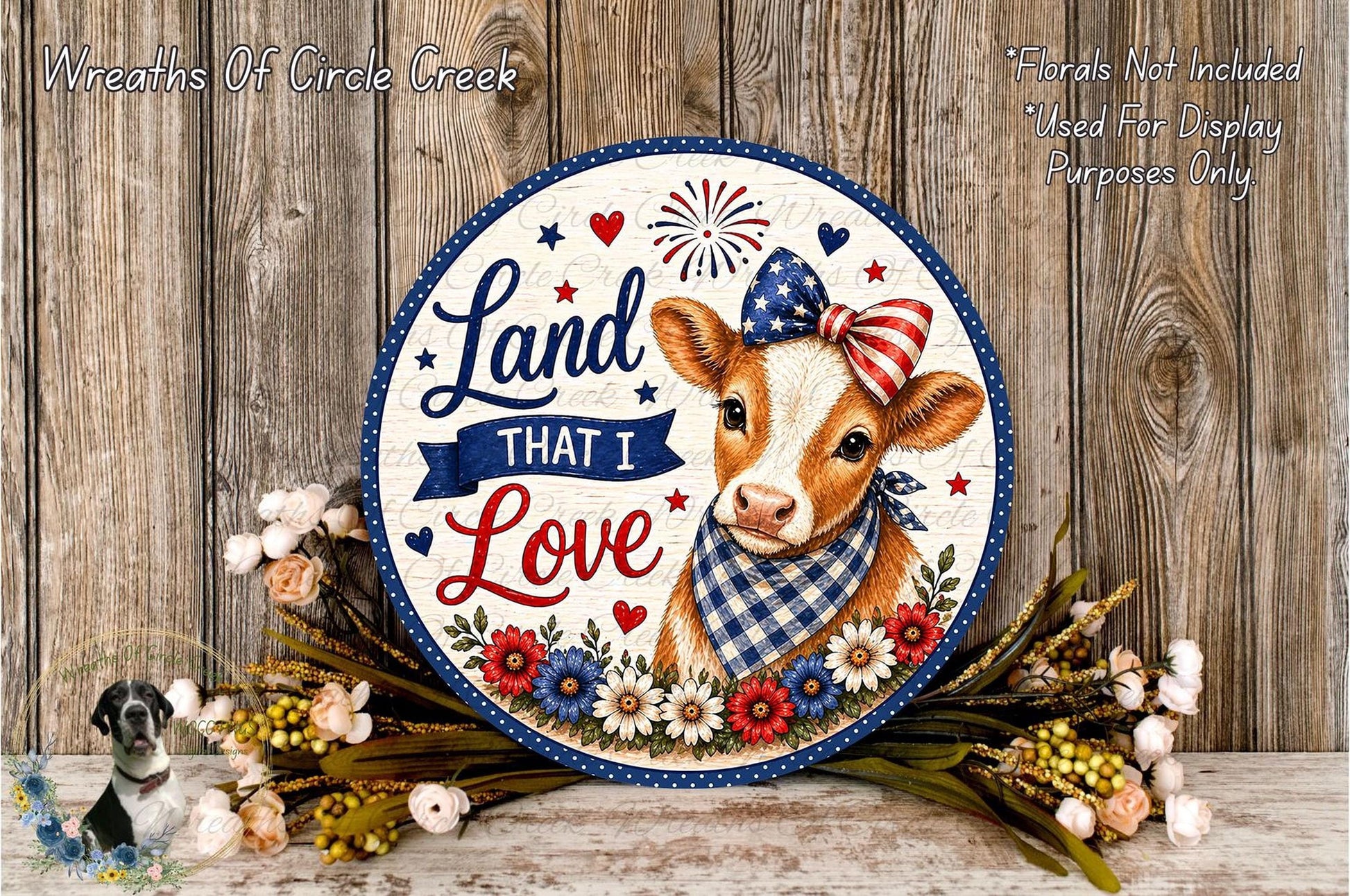 a decorative sign with a cow wearing a patriotic bow, surrounded by flowers and placed on a wooden surface.