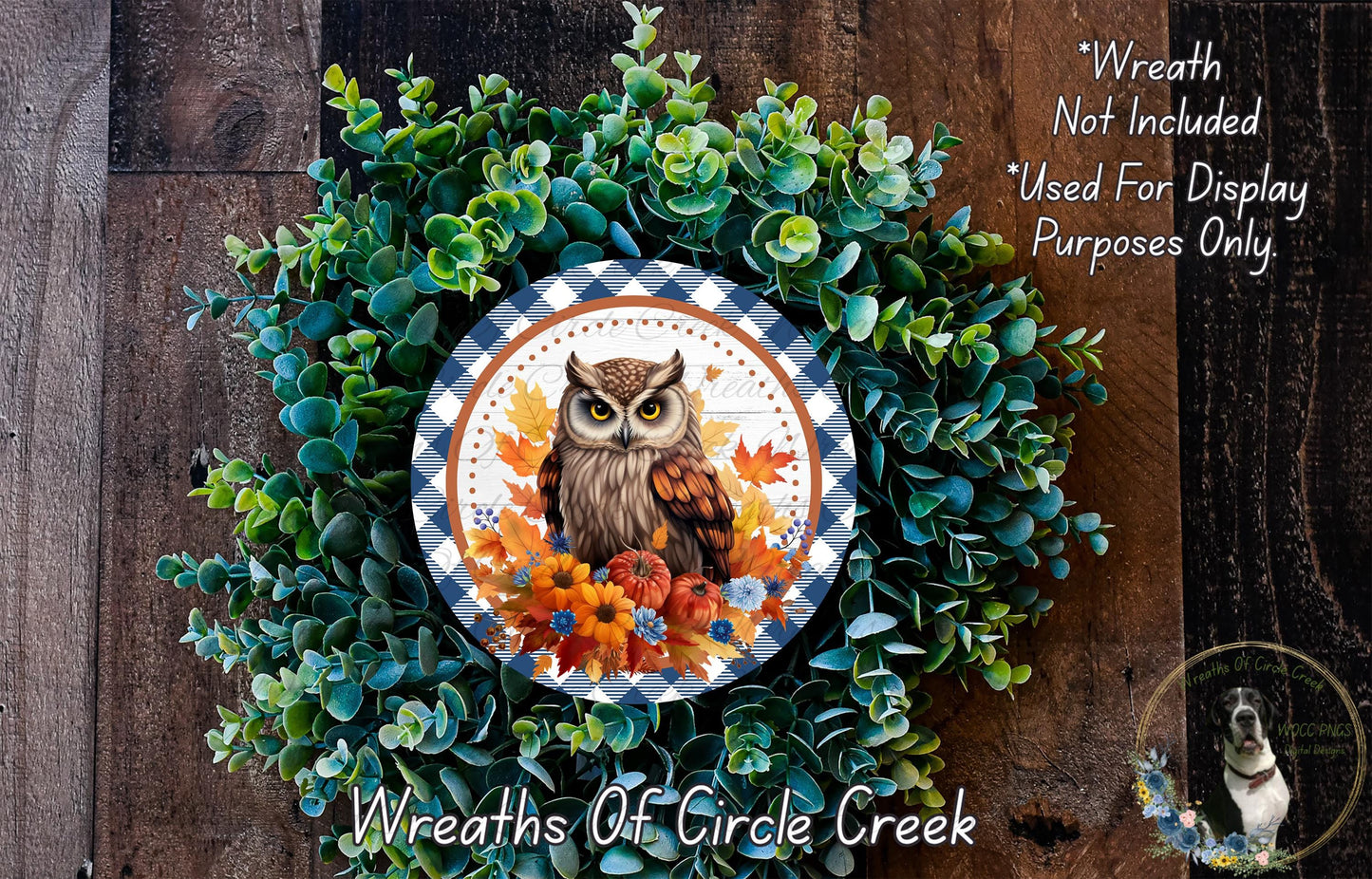 a wreath with an owl design, surrounded by green foliage and autumn leaves. The wreath is displayed on a wooden surface, and there is a small dog in the bottom right corner of the image.