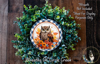 a wreath with an owl design, surrounded by green foliage and autumn leaves. The wreath is displayed on a wooden surface, and there is a small dog in the bottom right corner of the image.