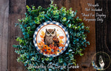 a wreath with an owl design, surrounded by green foliage and autumn leaves. The wreath is displayed on a wooden surface, and there is a small dog in the bottom right corner of the image.