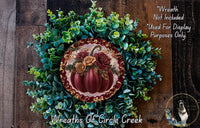 a wreath made of various green plants and a large red pumpkin with a gold border, placed on a wooden surface.