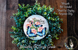 a wreath made of green leaves and flowers, with a circular design in the center that includes blue butterflies and pink flowers. The wreath is displayed on a wooden surface, and there is a small dog in the bottom right corner of the image.