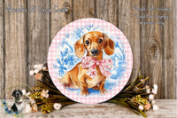 A round decorative plate featuring a dachshund dog wearing a pink bow, surrounded by a floral arrangement and placed on a wooden surface.