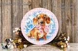 A round decorative plate featuring a dachshund dog wearing a pink bow, surrounded by a floral arrangement and placed on a wooden surface.