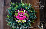 A whimsical wreath made of green leaves and flowers, featuring a large pink monster with a bow and green shoes, surrounded by a wooden background.
