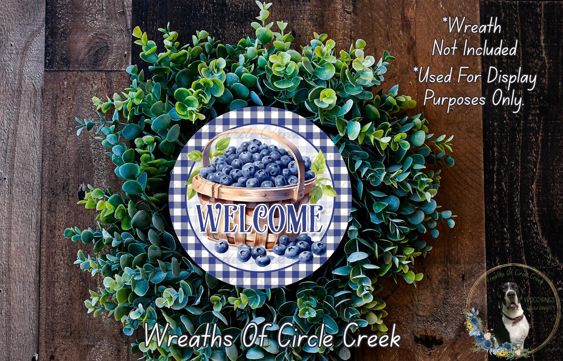 a wreath made of green leaves and blueberries, with a sign that reads "Welcome" in the center. The wreath is displayed on a wooden surface, and there is a small dog in the bottom right corner of the image.