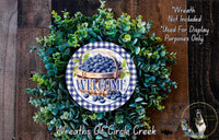 a wreath made of green leaves and blueberries, with a sign that reads "Welcome" in the center. The wreath is displayed on a wooden surface, and there is a small dog in the bottom right corner of the image.
