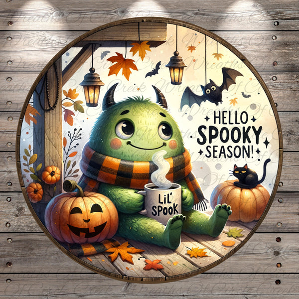 A whimsical illustration of a green monster sitting on a wooden surface, surrounded by autumn leaves, pumpkins, and bats, with a mug of what appears to be a hot beverage in front of it.
