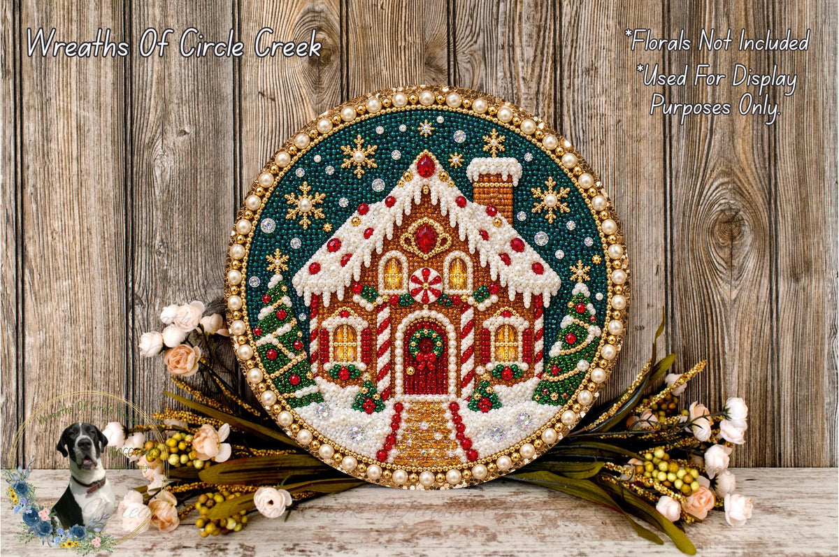 A decorative wreath featuring a gingerbread house, surrounded by flowers and placed on a wooden surface.