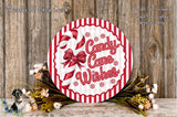 A round sign with the text "Candy Cane Wishes" and a candy cane decoration, surrounded by a wreath of flowers and greenery, placed on a wooden surface.