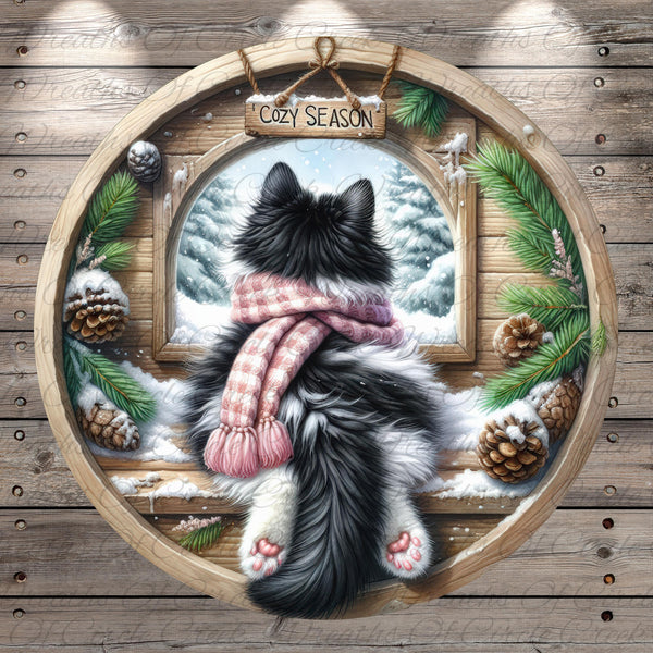 A black and white cat wearing a pink scarf is sitting on a wooden windowsill, looking out at a snowy winter landscape through a window.