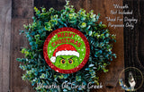 A Christmas wreath made of greenery, with a red and green circular ornament featuring a grumpy face wearing a Santa hat.