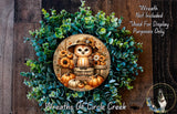 a wreath with an owl wearing a hat and surrounded by autumn-themed elements such as pumpkins and flowers. The wreath is displayed on a wooden surface, and there is a small dog in the bottom right corner of the image.