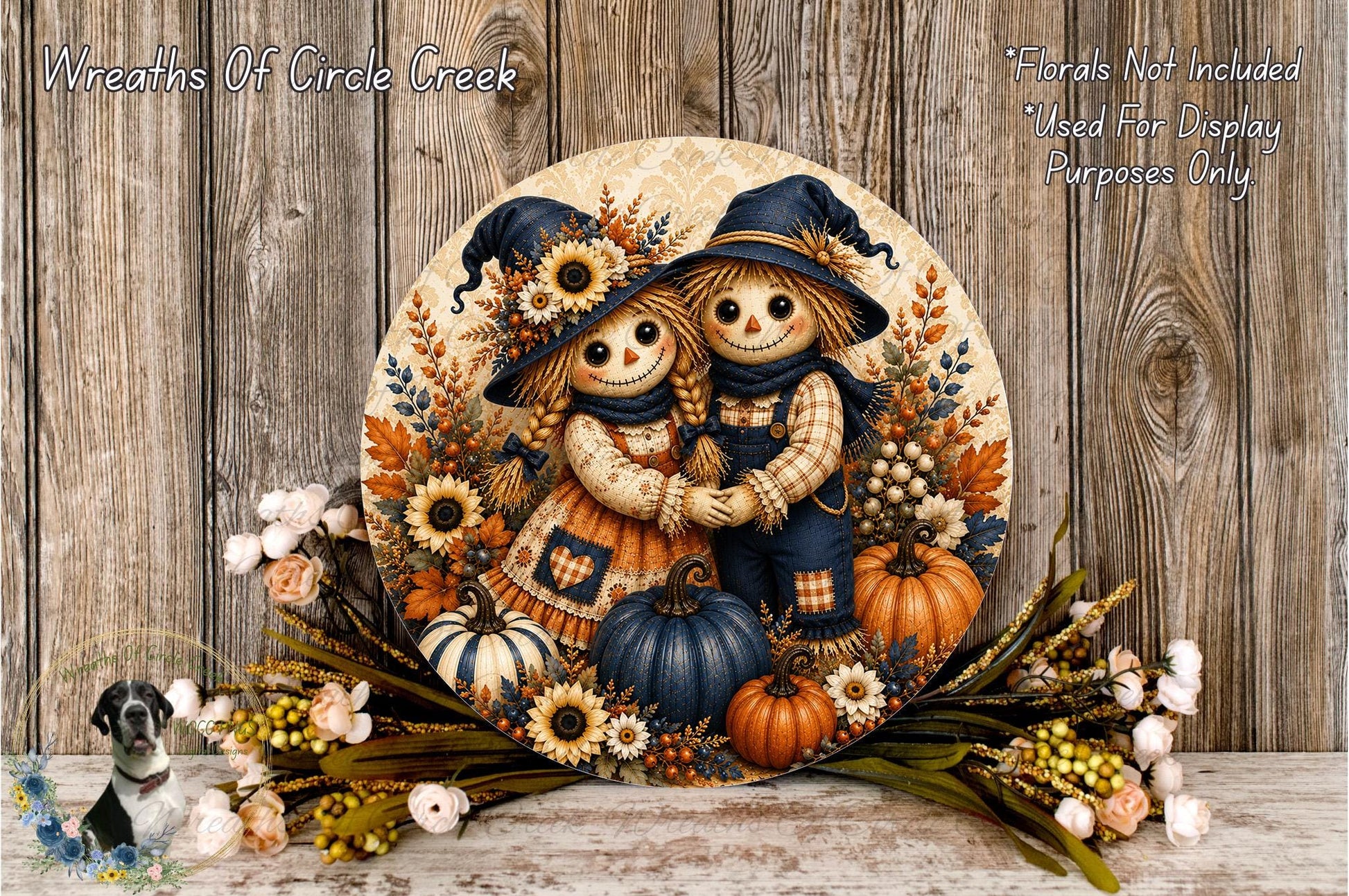A decorative wreath featuring two scarecrow dolls, surrounded by autumn foliage, pumpkins, and flowers, placed on a wooden surface.