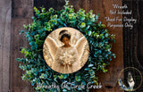 A wreath with a photo of a woman in a white dress and wings, surrounded by green leaves and flowers.