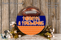A round sign with the text "Tailgates & Touchdowns" in orange and blue, surrounded by a wreath of flowers and a football.