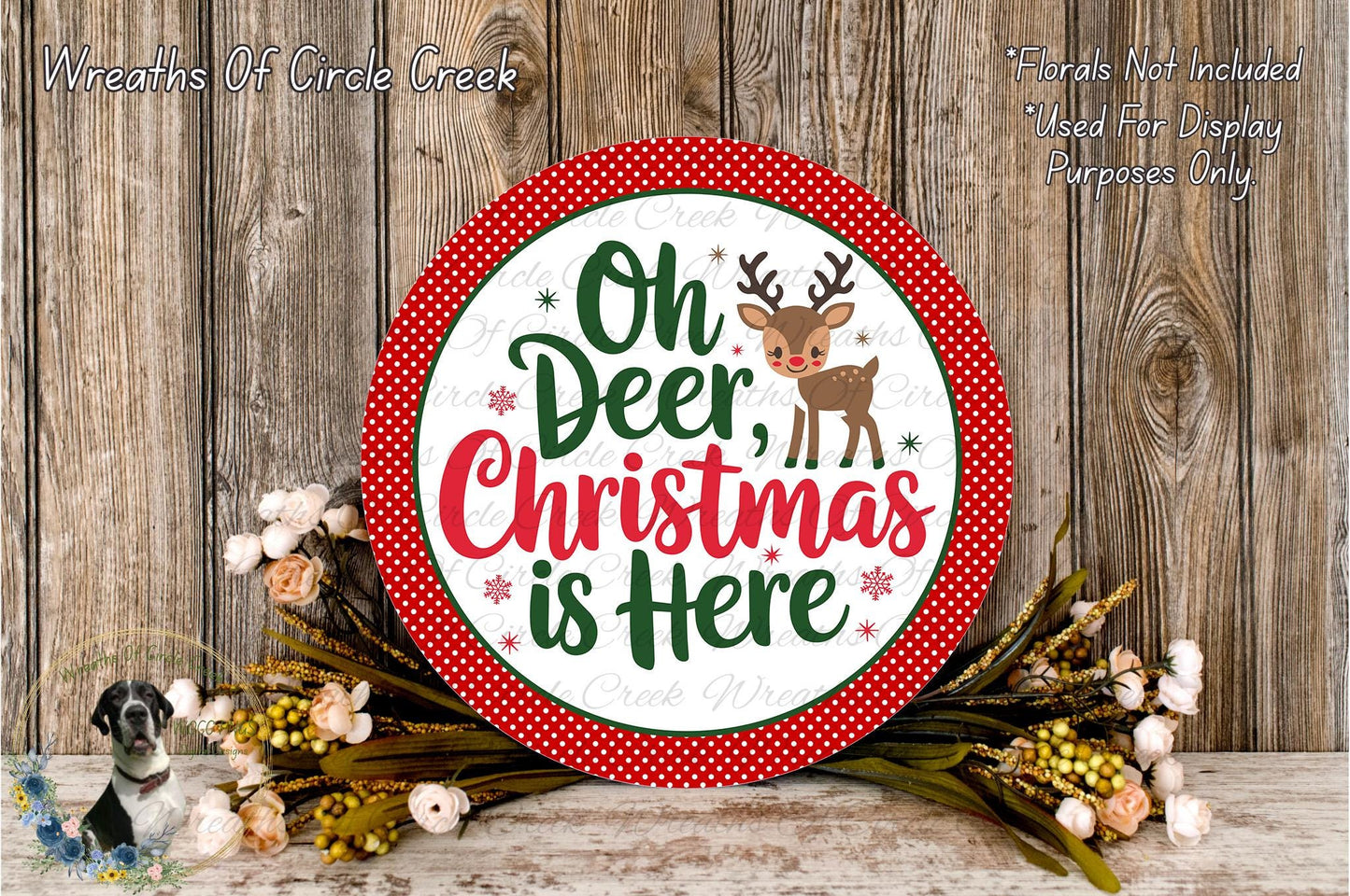 A circular sign with the text "Oh Deer, Christmas is Here" surrounded by a red border and a small reindeer illustration. The sign is placed on a wooden surface, with a wreath of flowers and greenery to the left and a wooden wall in the background.