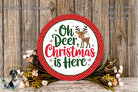 A circular sign with the text &quot;Oh Deer, Christmas is Here&quot; surrounded by a red border and a small reindeer illustration. The sign is placed on a wooden surface, with a wreath of flowers and greenery to the left and a wooden wall in the background.