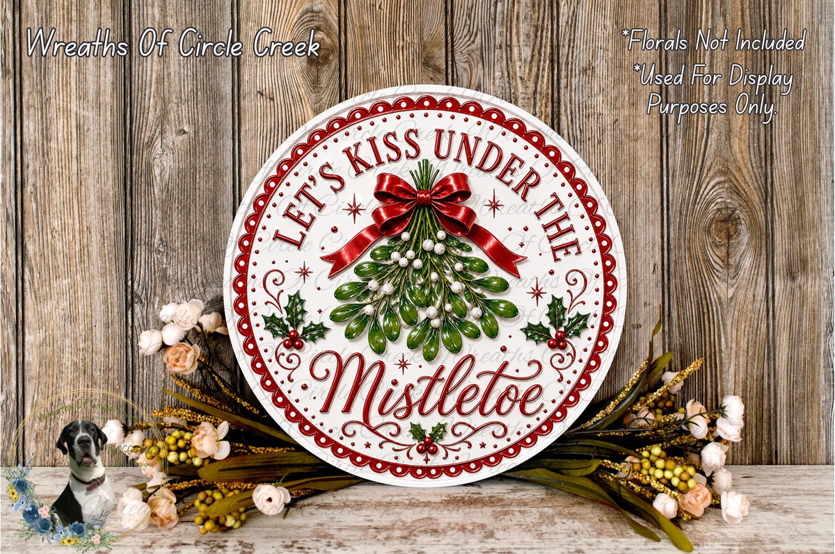 A decorative sign with the text "Let's Kiss Under The Mistletoe" surrounded by a wreath of greenery and flowers, placed on a wooden surface.