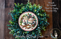 A wreath made of green leaves and other natural elements, with a decorative plate featuring a mug of hot chocolate and a snowman as the centerpiece.