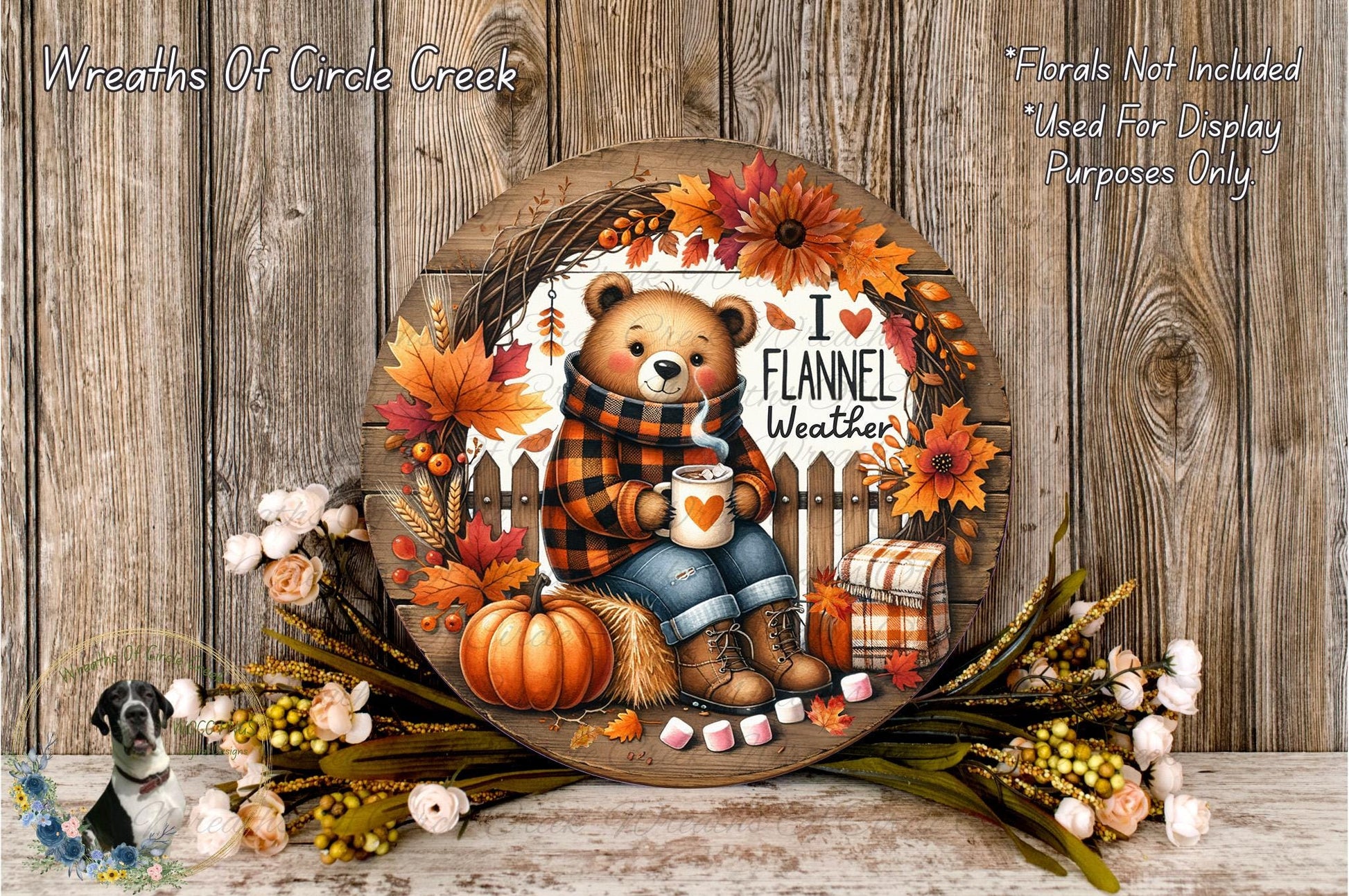 A wreath with a teddy bear wearing a plaid sweater and holding a cup of coffee, surrounded by autumn decorations such as pumpkins, leaves, and flowers, all set against a wooden background.
