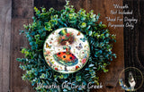 a wreath with a ladybug design, surrounded by green foliage and flowers. The wreath is displayed on a wooden surface, and there is a small dog in the bottom right corner of the image.
