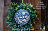 A wreath made of green leaves and a blue circular sign with the text "Christ the Savior is Born" displayed on a wooden background.