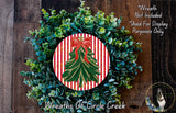 A Christmas wreath made of green and red evergreen branches, with a red bow on top, is displayed on a wooden surface.