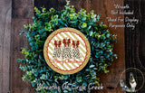 A Christmas wreath made of green leaves and a gold and red circular ornament with the text "Merry Christmas" on it, placed on a wooden surface.