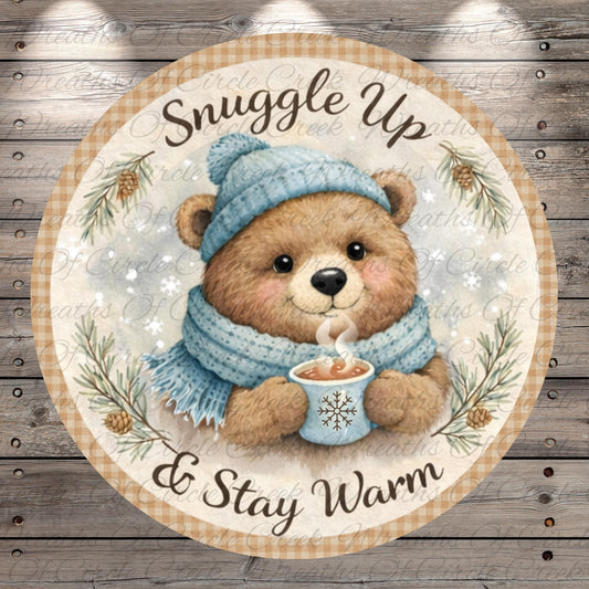 A round sticker with a teddy bear wearing a blue hat and scarf, holding a cup of hot chocolate, and surrounded by pine branches and snowflakes. The sticker has the text "Snuggle Up & Stay Warm" written on it.