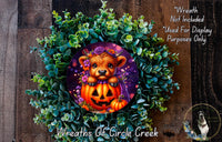 A wreath made of green plants and a pumpkin with a carved face, featuring a small brown bear wearing a purple hat.