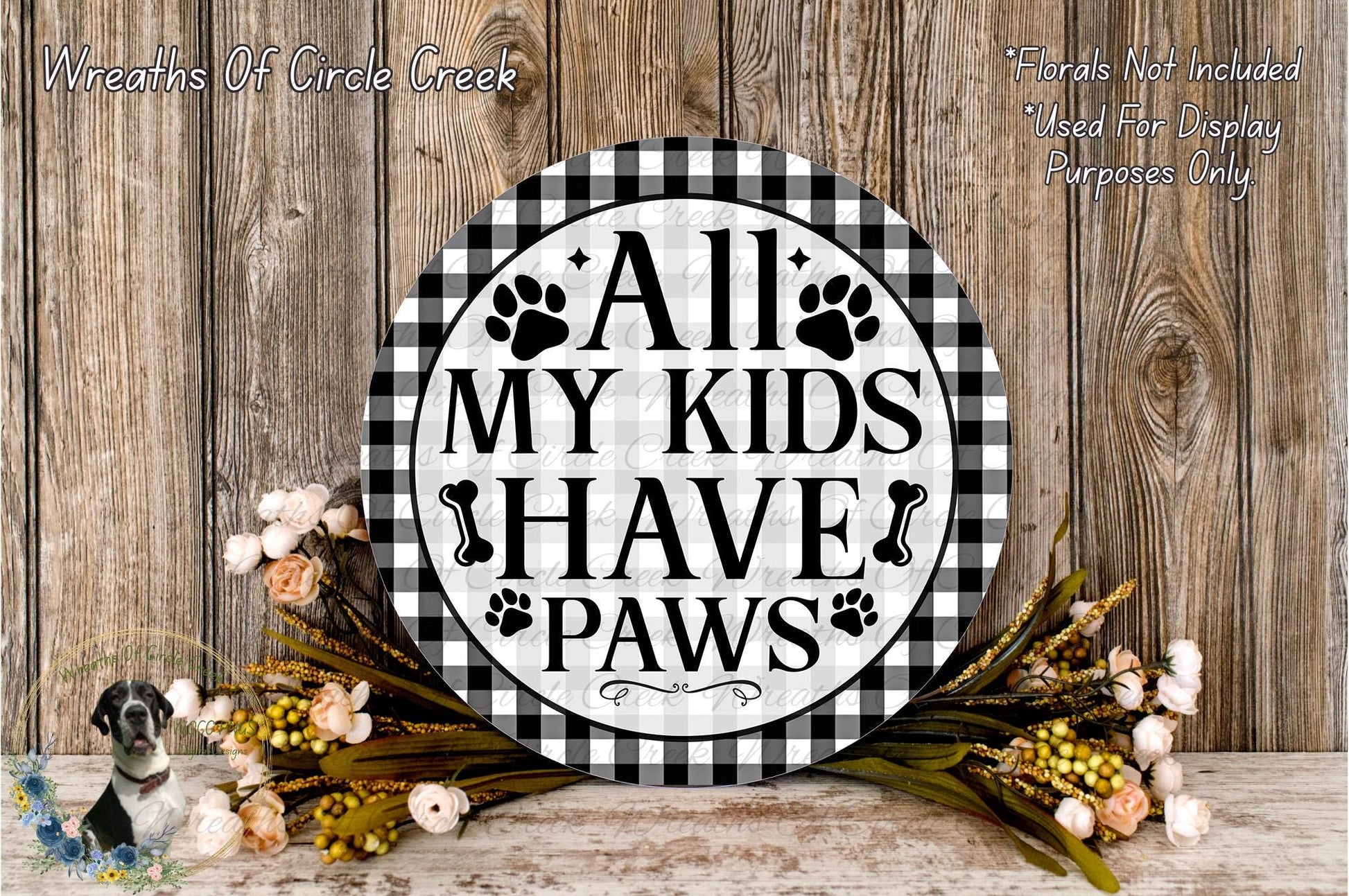 a decorative sign with the text "All My Kids Have Paws" displayed on a wooden surface. The sign is surrounded by a floral arrangement, and there is text in the top left corner that reads "Wreaths Of Circle Creek".