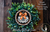 a wreath made of green leaves and a black and white pumpkin with an orange bow, displayed on a wooden surface.
