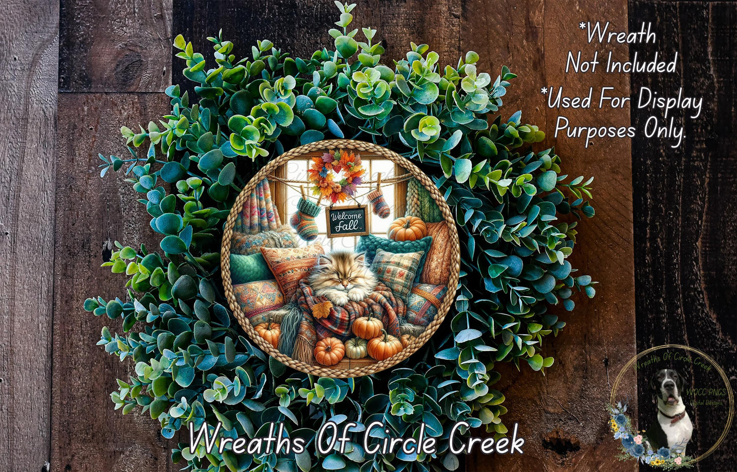 A wreath made of green leaves and pumpkins, with a cat sitting on a pillow inside the wreath. The wreath is displayed on a wooden wall.
