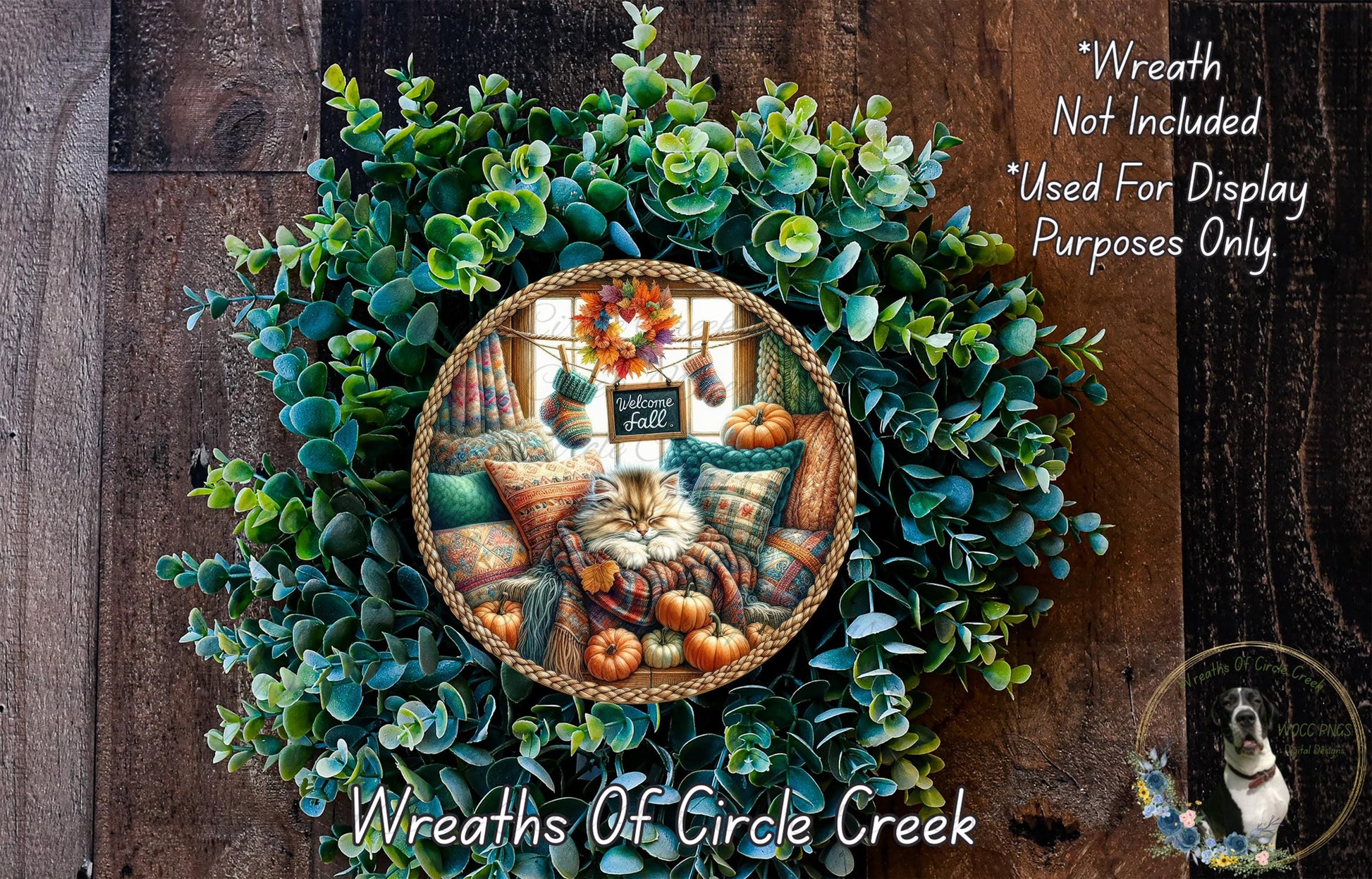 A wreath made of green leaves and pumpkins, with a cat sitting on a pillow inside the wreath. The wreath is displayed on a wooden wall.