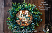A wreath made of green leaves and pumpkins, with a cat sitting on a pillow inside the wreath. The wreath is displayed on a wooden wall.