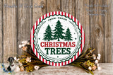 a circular sign with the text "Christmas Trees" surrounded by a wreath of greenery and flowers, set against a wooden background.